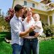 Family of three smiling in front of a house with a green lawn and plants.