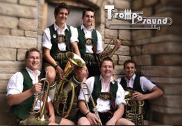 Fünf Männer in traditioneller Kleidung mit Blasinstrumenten vor einer Steinmauer.