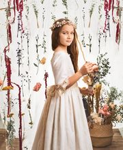 Niña con vestido blanco y corona de flores, rodeada de decoraciones florales colgantes.