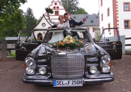 Brautpaar jubelt aus einem Oldtimer mit Blumenschmuck vor historischer Kulisse.