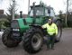 Eine Person in Warnweste steht neben einem großen grünen Traktor auf einem gepflasterten Weg.