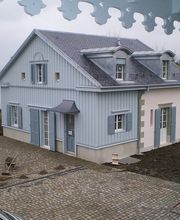 Ein graues Haus am Wasser, umgeben von Pflastersteinen und einer Rasenfläche, grauer Himmel.