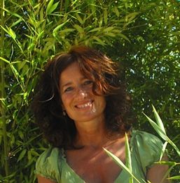 Eine lächelnde Frau in einem grünen Oberteil steht vor vielen Pflanzen.