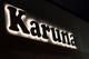 Letrero iluminado con la palabra "Karuna" en letras grandes y oscuras sobre fondo luminoso.