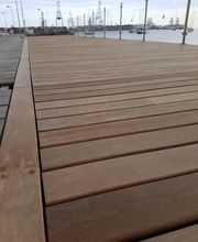 Muelle de madera junto al agua con grúas y barcos en el horizonte bajo un cielo nublado.