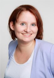 Frau mit kurzen, roten Haaren lächelt in die Kamera; trägt hellblauen Blazer und weißes Shirt.