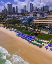 Praia com guarda-sóis azuis, prédios altos ao fundo e mar à esquerda sob céu azul.
