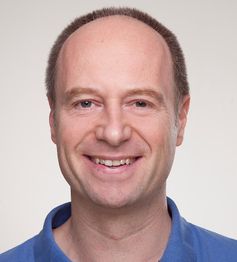 Ein lächelnder Mann in einem blauen Hemd vor einem neutralen Hintergrund.