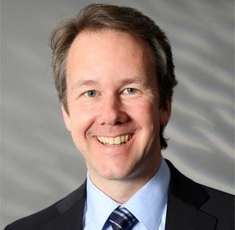 Ein lächelnder Mann in Anzug und Krawatte vor einem grauen Hintergrund.