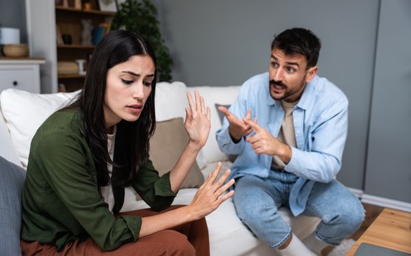 Ein junges Paar sitzt auf dem Sofa und streite sich. Der Mann macht der Frau gestikulierende Vorwürfe.