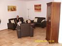 Wohnzimmer mit Couchgarnitur, Lampe, Vitrinenschrank und Wandbildern. Zwei Personen sitzen.