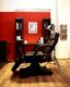 Barbershop-Stuhl vor roter Wand, flankiert von Regalen und Lesematerialien, Lampenbeleuchtung.
