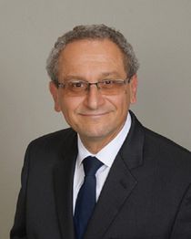 Smiling man in a suit with glasses and curly hair against a gray background.