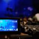 Close-up of a video camera recording a dimly lit, blue-toned stage performance.