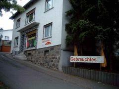 Laden in einem weißen Gebäude mit einem Schild "Gebrauchtes" an einer leicht ansteigenden Straße.