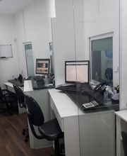 Escritório com cadeiras, computadores em mesas brancas e divisórias de vidro. Piso de madeira.