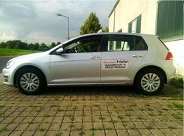 Silberner Pkw auf gepflastertem Parkplatz vor Gebäude, mit Werbeaufkleber auf der Tür.