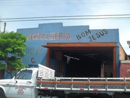 Caminhão branco estacionado com uma serralheria "Bom Jesus" ao fundo, fachada azul e letreiro.