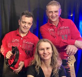 Eine Frau sitzt; zwei Männer in roten Hemden stehen daneben. Einer hält ein Tamburin, der andere eine Gitarre.