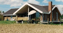 Ein Backsteinhaus mit Satteldach und Terrasse steht vor einem Feld unter blauem Himmel.
