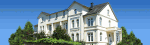 Weißes Herrenhaus mit Dachgauben und Garten, vor blauem Himmel.