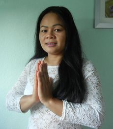 Frau in weißem Spitzenoberteil mit gefalteten Händen vor einer mintgrünen Wand.