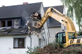 Excavator demolishing a house, breaking through the roof and walls.