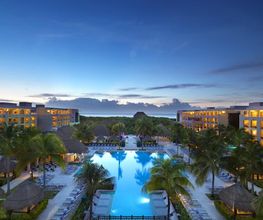 Complejo turístico con piscina al atardecer, rodeado de palmeras y edificios iluminados.