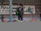Eine Person spielt Gitarre auf einer Bühne vor einem Schild und einem Mikrophon.