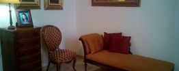 Sala con chaise longue, silla, cómoda, lámpara y fotos en marcos dorados. Cojines rojos.