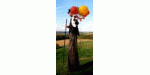 Person auf Stelzen hält bunte Luftballons, steht auf grüner Wiese mit blauem Himmel.