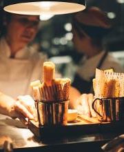 Chefs de cozinha preparando prato com copos de batatas fritas, em ambiente iluminado.