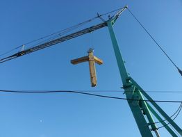 Una croce di legno viene sollevata da una gru contro un cielo blu.