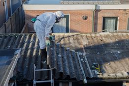 Person in protective gear removing roofing materials on a ladder.