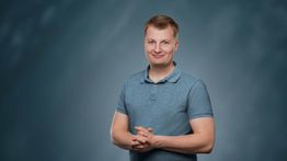 Eine lächelnde Person in einem blauen Polohemd steht vor einem grauen Hintergrund.