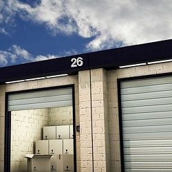 Storage unit 26 with open door, stacked with cardboard boxes. Cloudy blue sky above.
