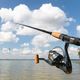 Fishing rod with reel near a lake, under a blue sky with fluffy clouds.