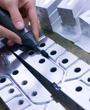 Hände arbeiten mit einem Werkzeug an einer Metallform; technische Zeichnungen im Hintergrund.