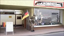 Schaufenster eines Waffengeschäfts, Soldatenfigur mit deutscher Flagge davor. Schild über dem Laden.