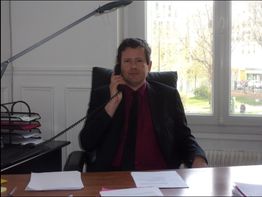 Homme assis à un bureau, téléphonant, avec des papiers devant lui et des dossiers empilés.