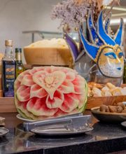 Bufê decorado com melancia esculpida, máscara veneziana e azeite, ao fundo pães e queijos.