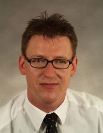 Mann mit Brille, weißem Hemd und Krawatte vor neutralem Hintergrund.