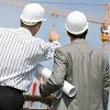 Two engineers in hard hats review construction site plans with cranes in the background.