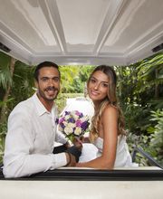 Pareja sonriente en un carro de golf, ella sostiene un ramo de flores. Fondo de vegetación tropical.