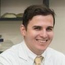 Pessoa sorrindo, vestindo jaleco branco e gravata amarela, em ambiente interno com prateleiras ao fundo.