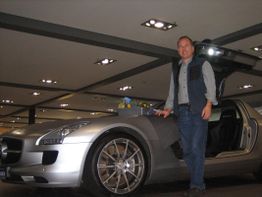 Ein Mann steht neben einem silbernen Auto mit geöffneten Flügeltüren in einem Ausstellungsraum.