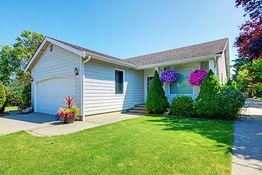 White house with a garage, green lawn, and colorful flowers hanging by the window.
