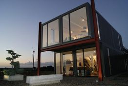 Modernes zweistöckiges Containerhaus bei Sonnenuntergang mit großem Fenster und Windrad im Hintergrund.