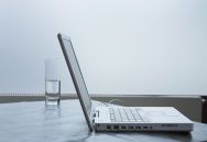 Laptop und Glas Wasser auf einem Tisch vor neutralem Hintergrund.
