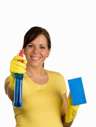 Frau in gelbem T-Shirt und Handschuhen hält Reinigungsmittel und blauen Schwamm, lächelnd.